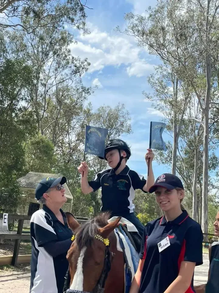 Boy on horse holds flags, two women stand nearby, trees and sky in background.
