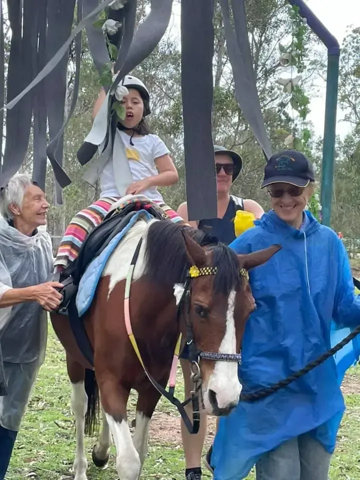 Child rides horse, led by adults. Horse is brown and white. Outdoors with streamers.