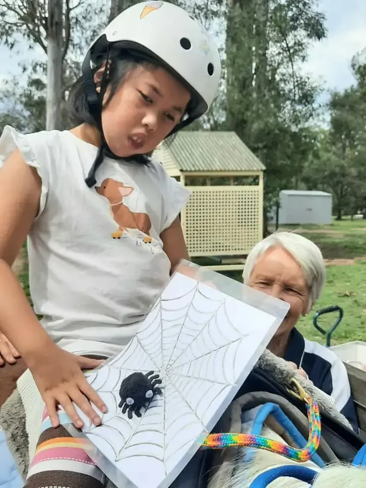 Girl in helmet holds spider web drawing with a woman in a park.