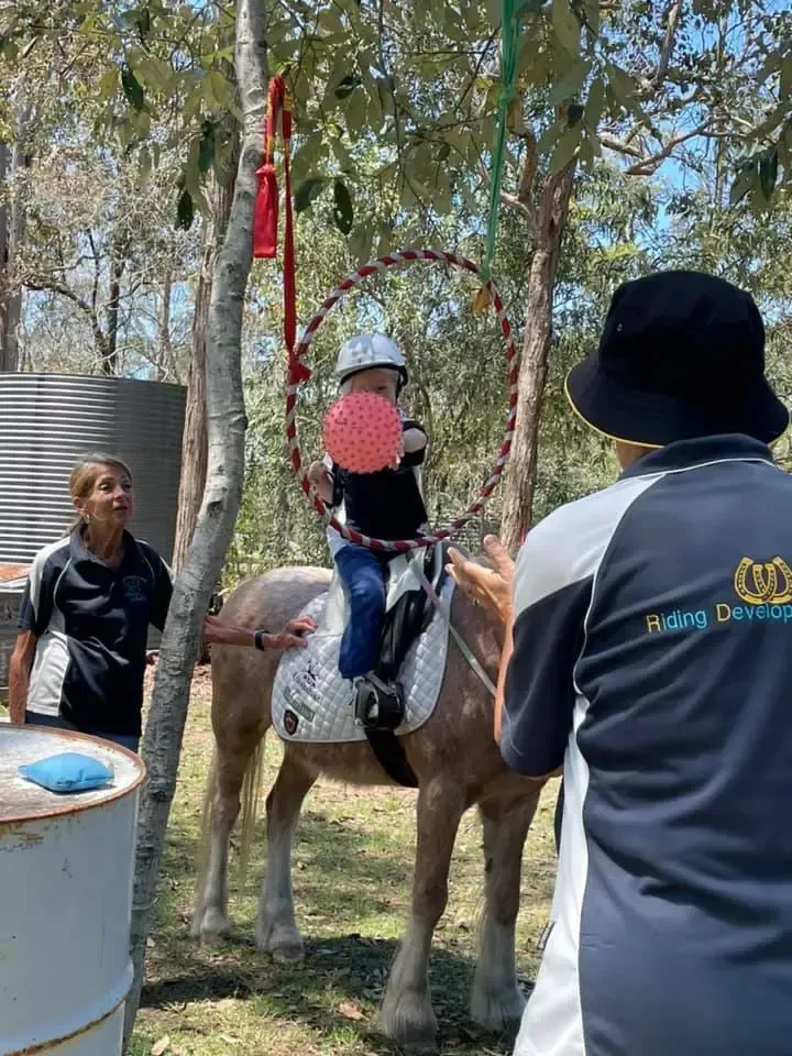 Child on horse reaching for ball through hoop, assisted by two adults in outdoor setting.