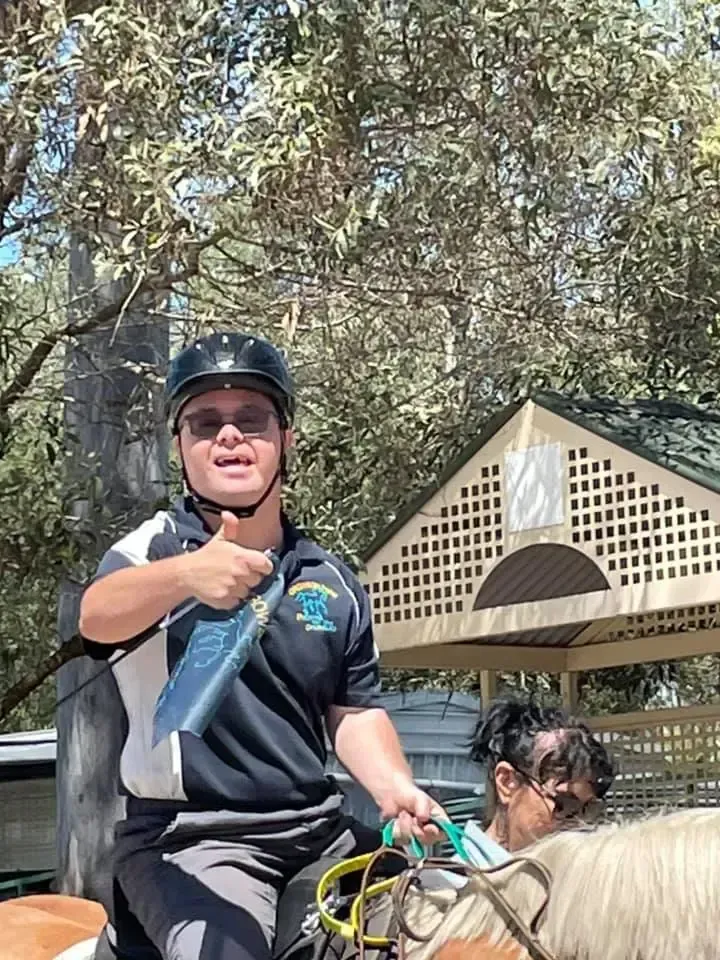 Person on horseback giving a thumbs-up, wearing a helmet. Outside, near a structure.