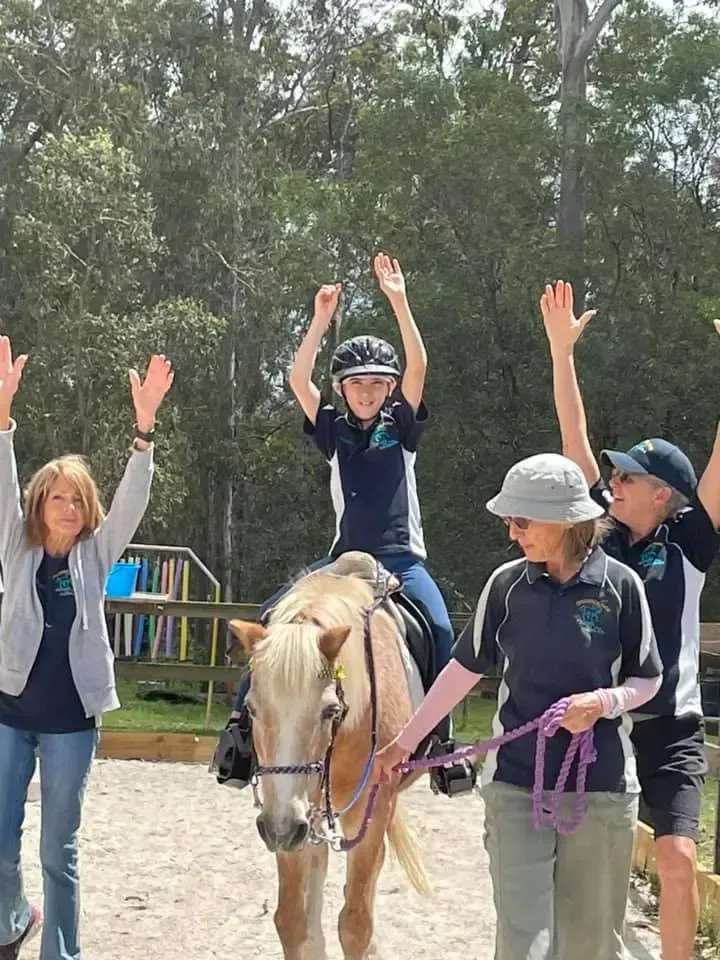 Child on horse with arms raised, surrounded by adults also raising arms; outdoors, sunny.