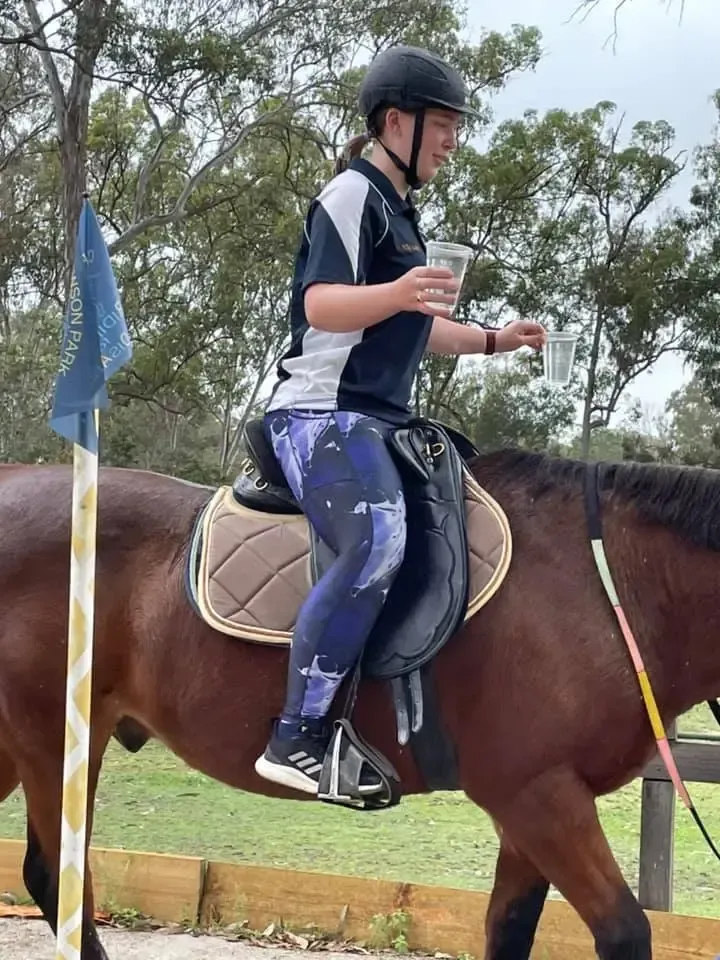 Person on a brown horse, wearing helmet and riding attire. Holding a cup. Outdoors, near a fence.