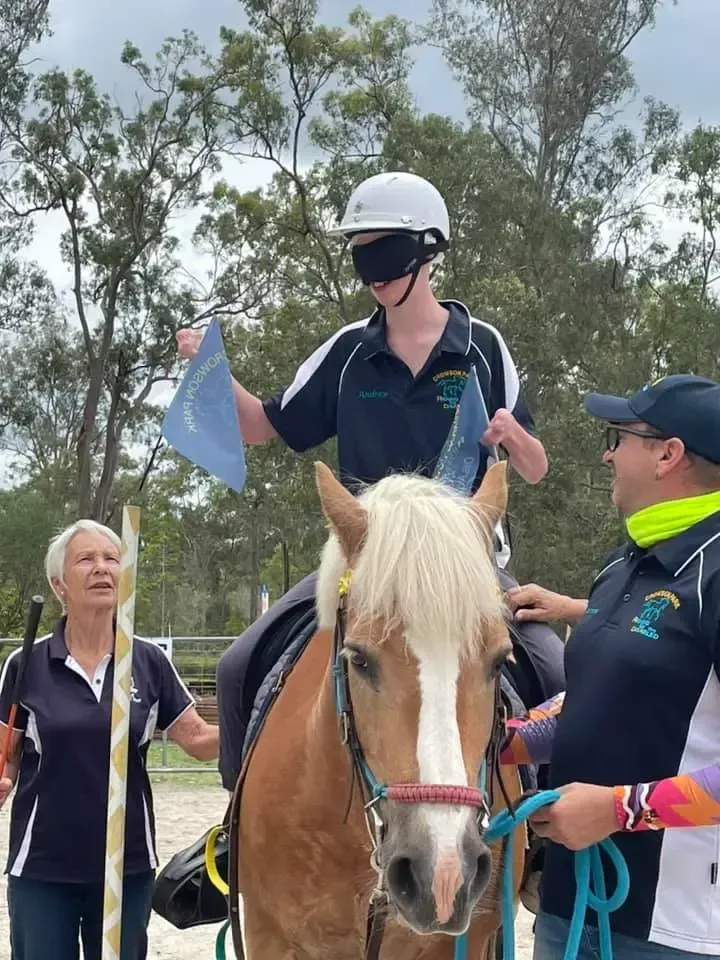 A person with a blindfold rides a horse, holding flags. Two people assist. Outdoor setting.