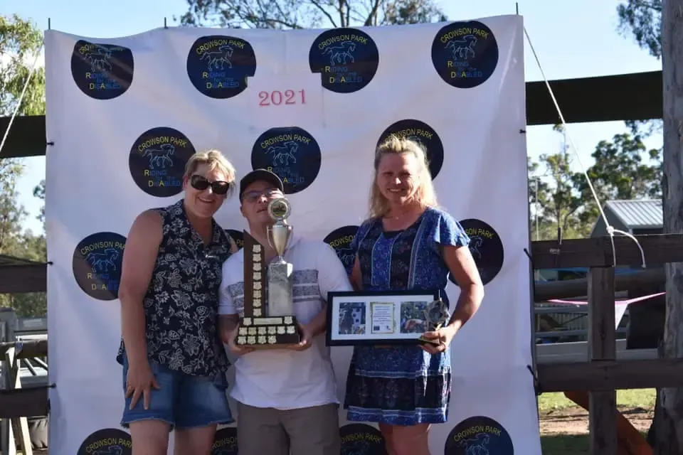 Three people smiling, holding a trophy and framed photos in front of a banner.