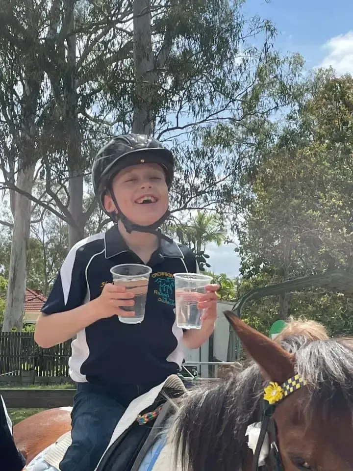 Boy on horseback smiles, holding two clear cups. Outdoors, trees in background, horse has yellow flower.