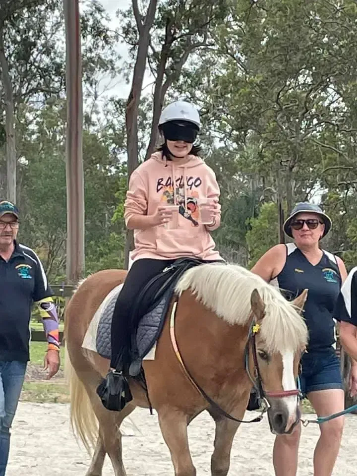 Person riding a pony with blindfold, guided by adults, outdoor setting.