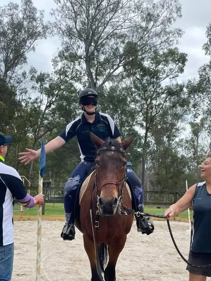 Person on horseback with blindfold, arms outstretched; two spotters.