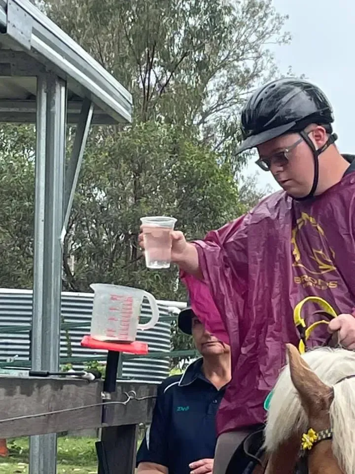 Person on horseback, wearing rain gear, holding a cup near a measuring container. Another person observes.
