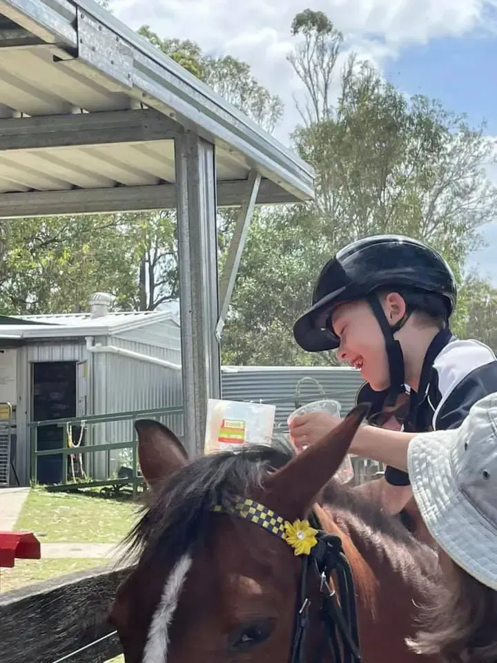 Boy in riding helmet on a brown horse, petting it. Outdoor setting with a shed in the background.
