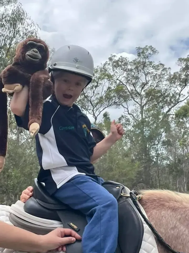 Boy on horse, wearing a helmet, holding a stuffed monkey, smiling and giving a thumbs-up gesture.