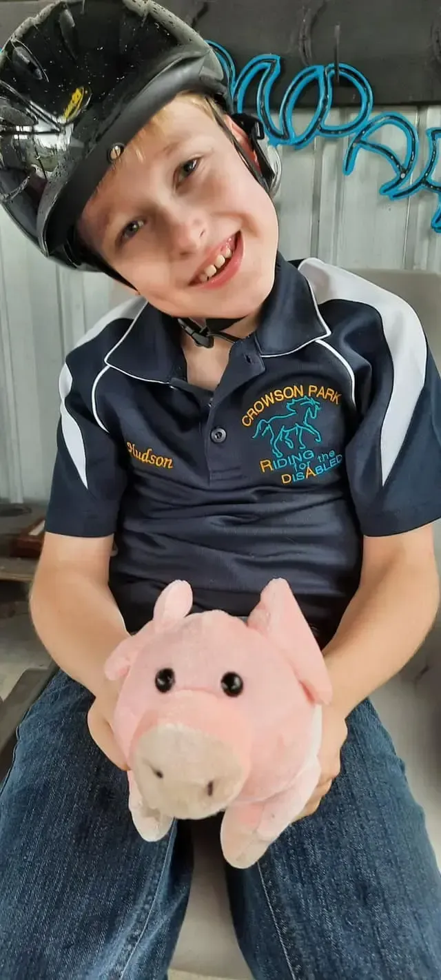 Boy in helmet smiles, holding pink pig plushie. Blue shirt, denim jeans.
