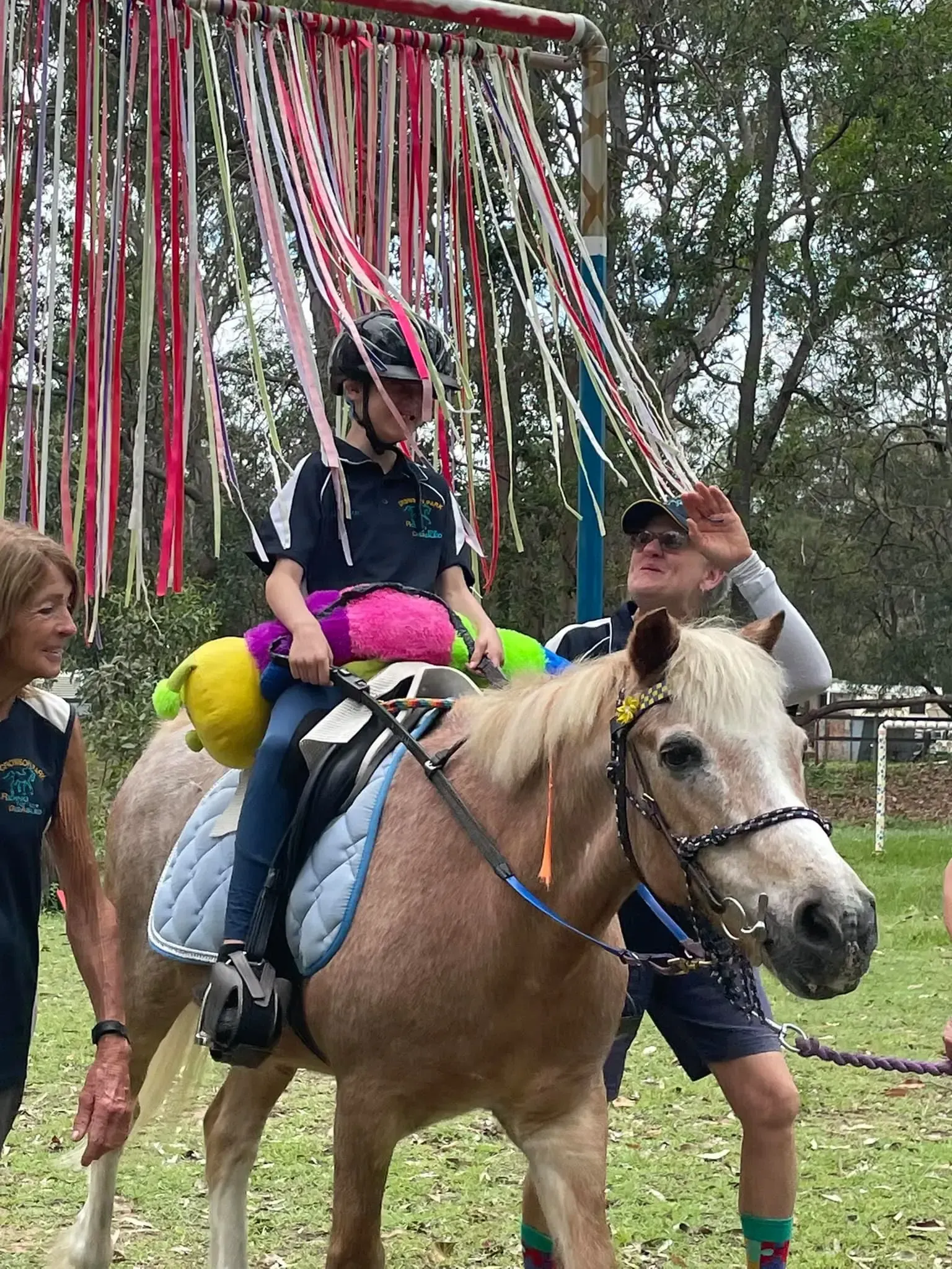 Child rides a pony through a ribbon archway, guided by two adults. Outdoor setting.