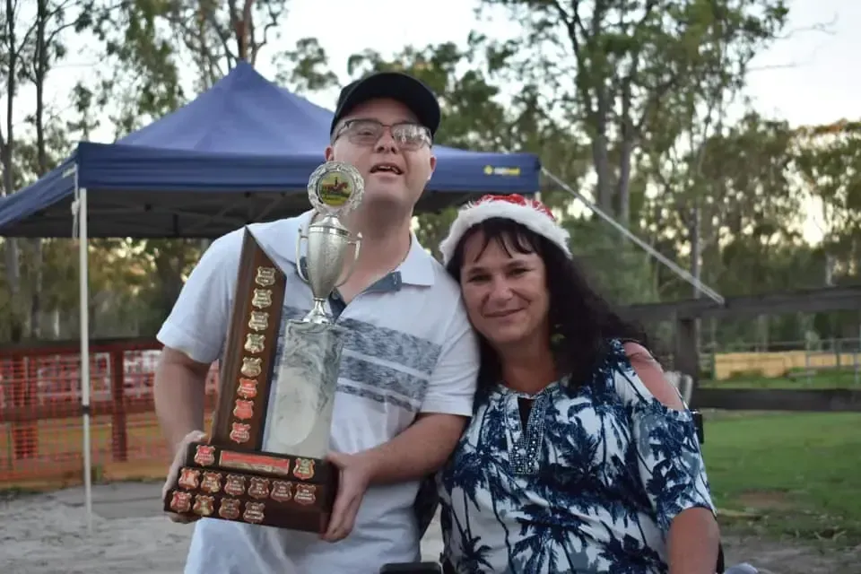 A smiling person with a large trophy poses with a woman wearing a Santa hat under a blue canopy.