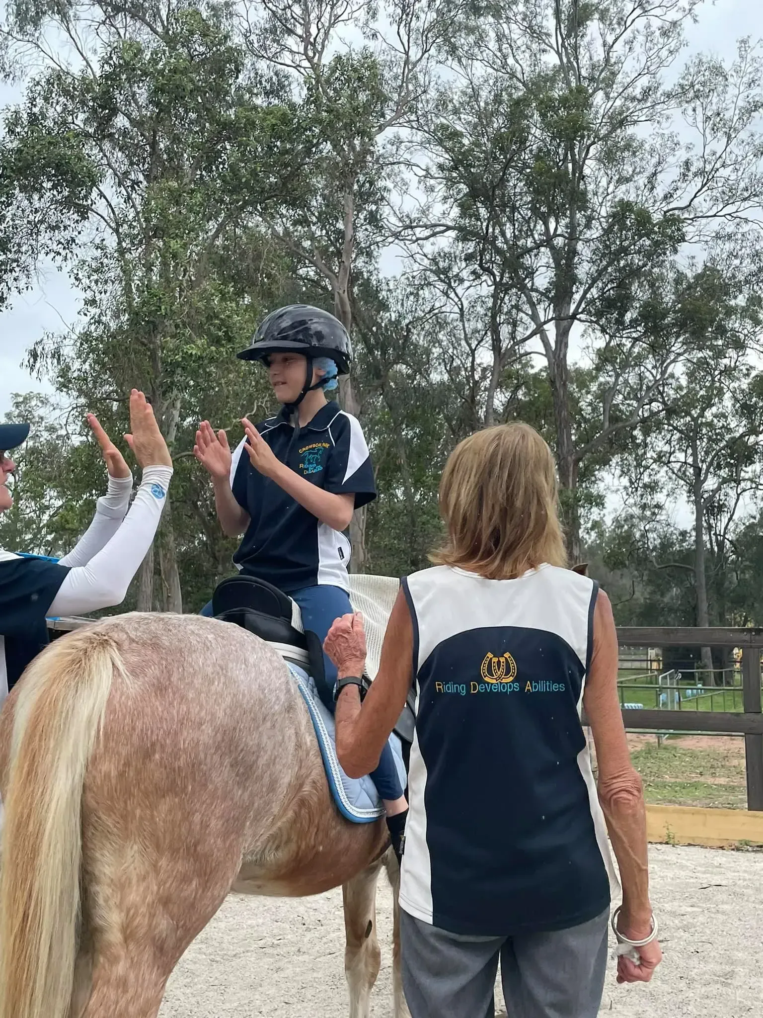 A child in a riding helmet claps hands while on a horse; two adults cheer, one giving a high five. Outdoors.