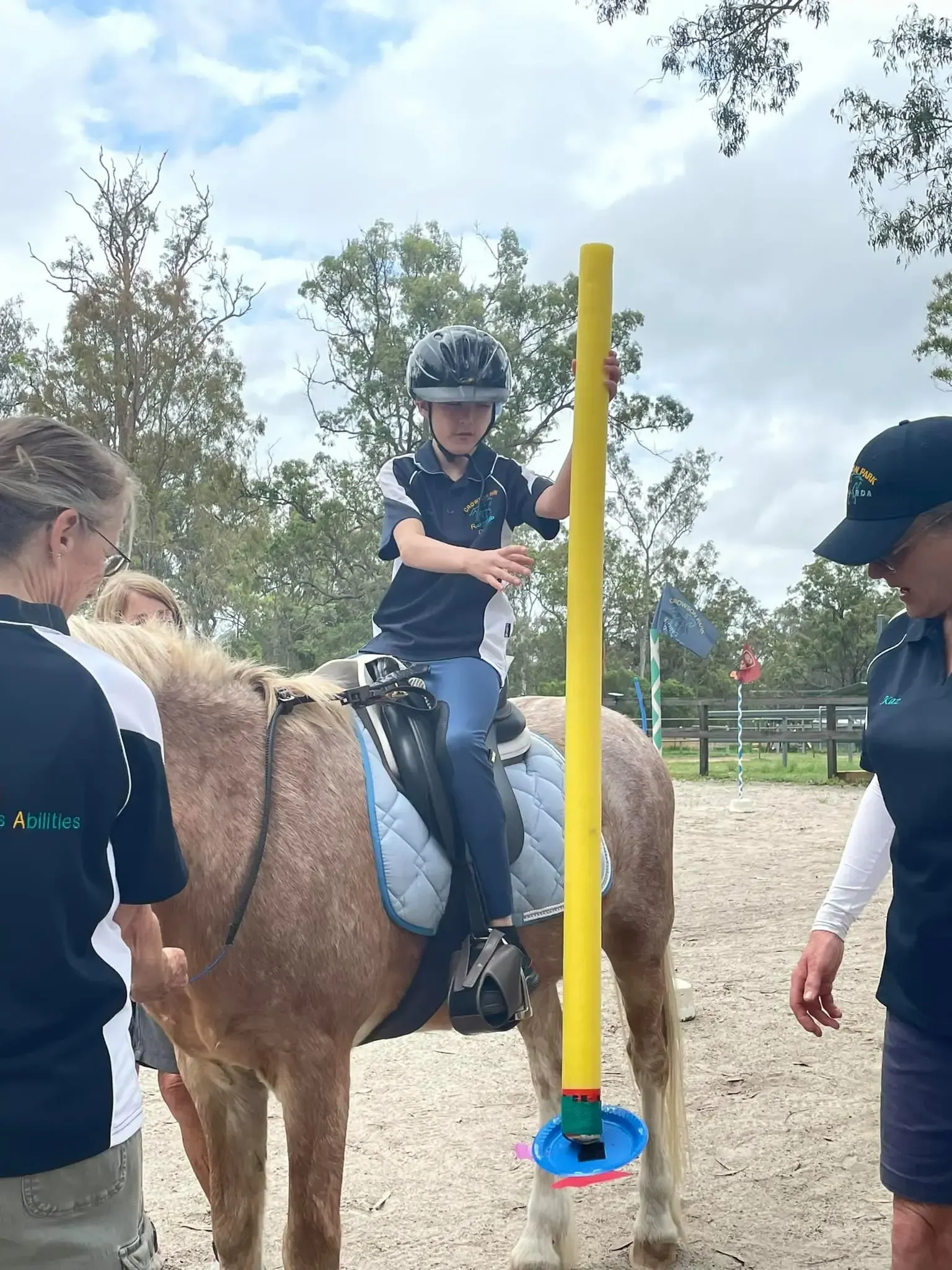 A child on a pony holds a yellow pole during an equestrian lesson. Two adults stand nearby.