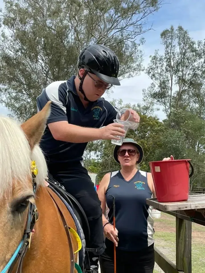 Person on horseback pours water into a bucket; a woman with a riding crop watches. Outdoor setting.
