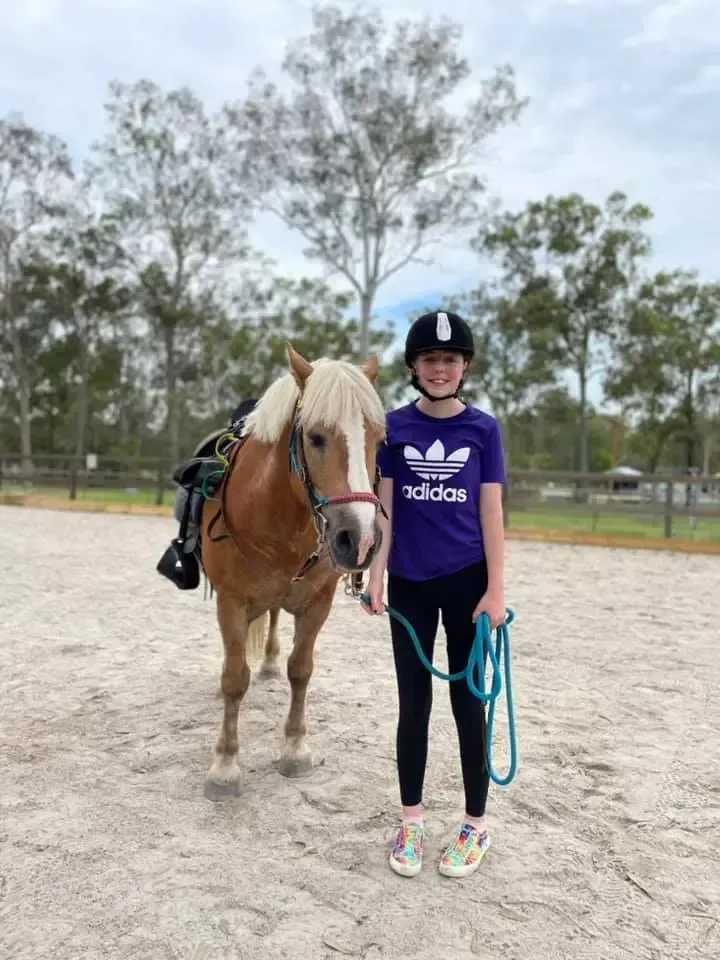 Girl in riding helmet with pony in a sandy arena. Pony is light brown with a blonde mane.