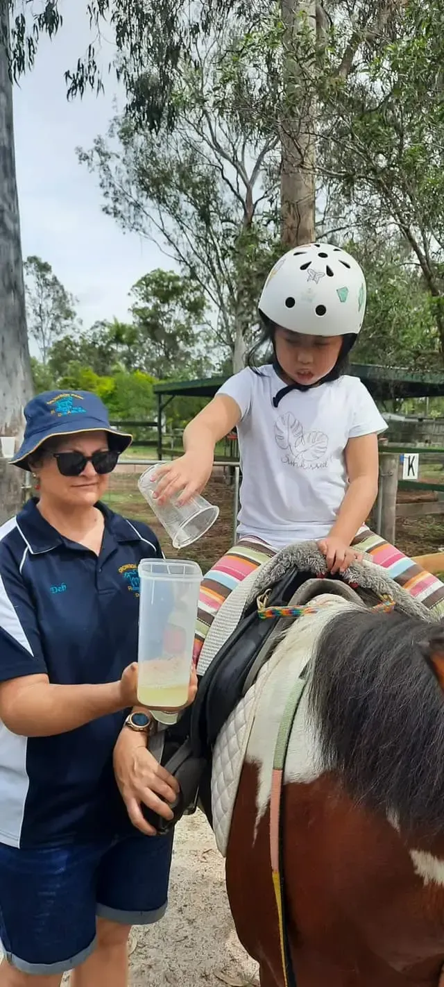 A child on a pony pouring liquid from a cup into a bottle held by an adult. Outdoors.