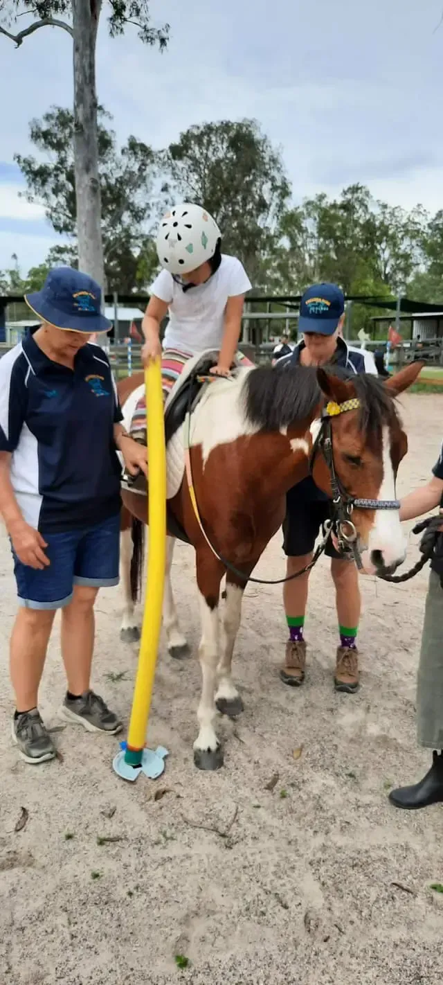 A child rides a horse with help from adults. A yellow pole stabilizes the child, outdoors on a sandy surface.