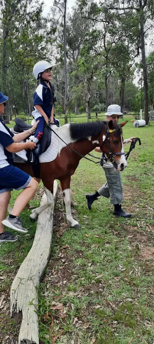 A child on horseback is led over a log obstacle. Two people assist, and a third holds the horse's reins.