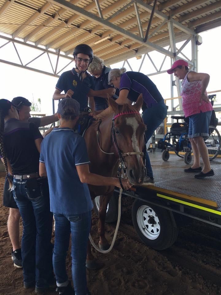 People assisting a horse onto a trailer ramp. Several people touch the horse. A person in a wheelchair watches.