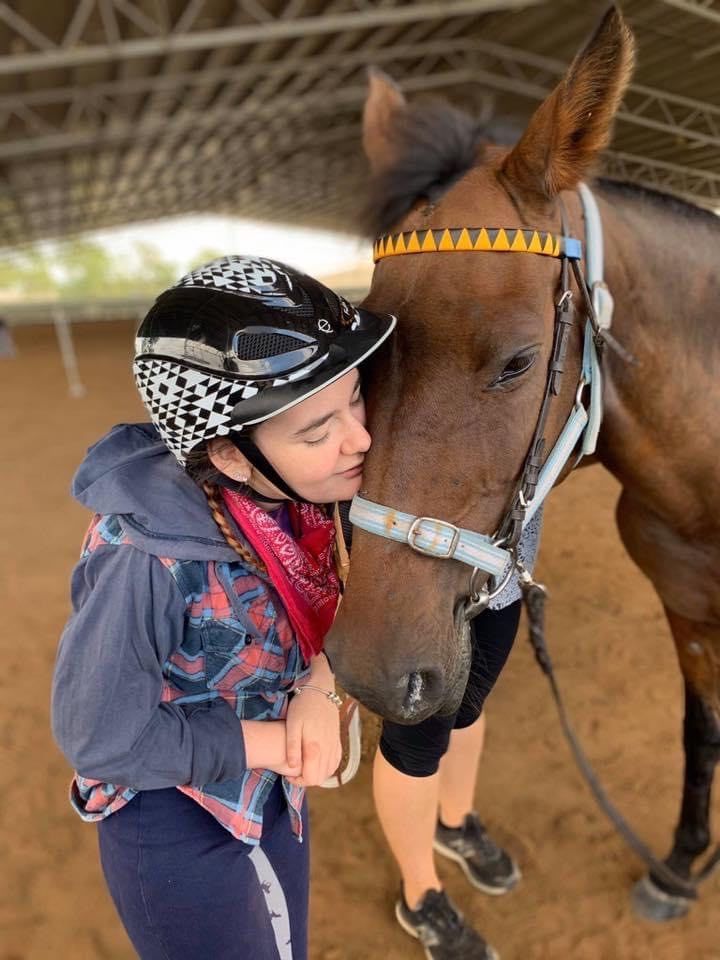 Person wearing a helmet hugs a brown horse. They're indoors under a canopy with dirt floor.