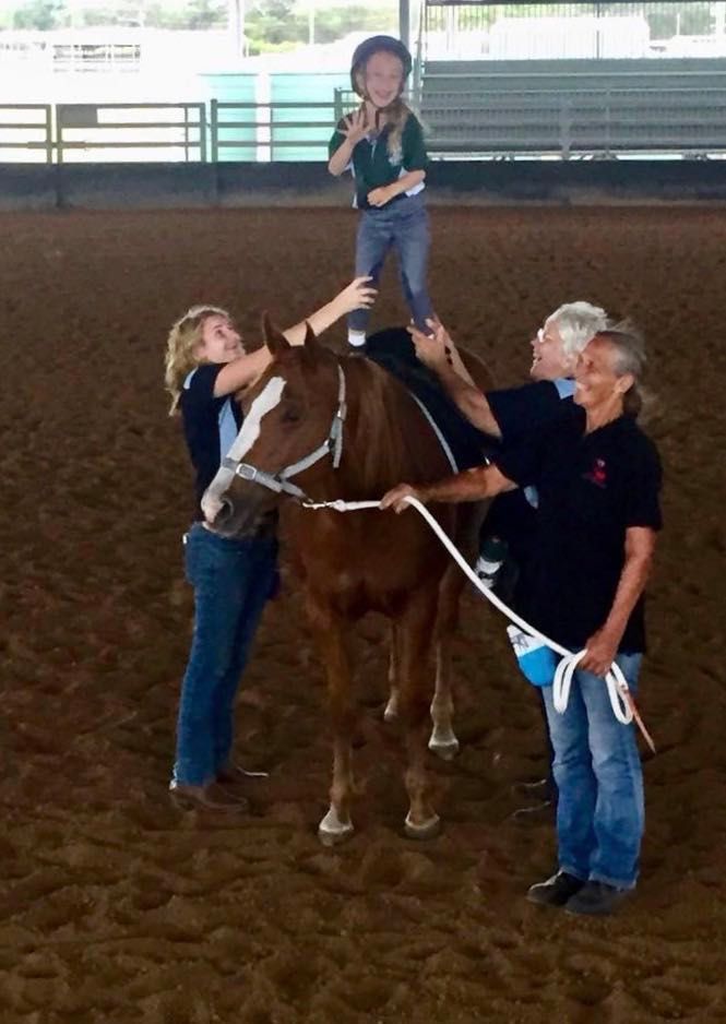 Girl on horse, helped by three adults in indoor arena. Child waves, others hold and steady horse.