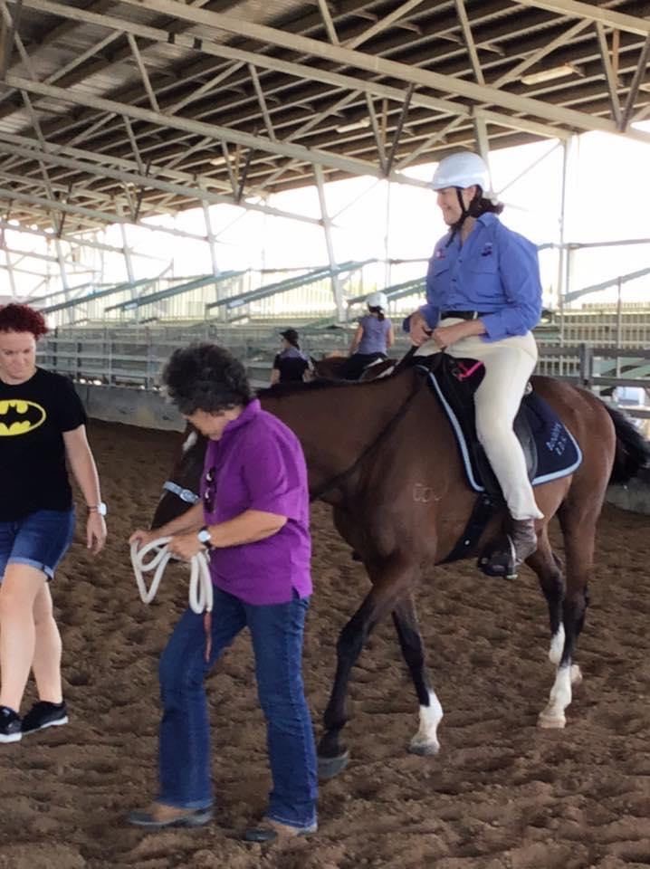 Woman riding horse in an indoor arena, two others assisting; one holding reins.