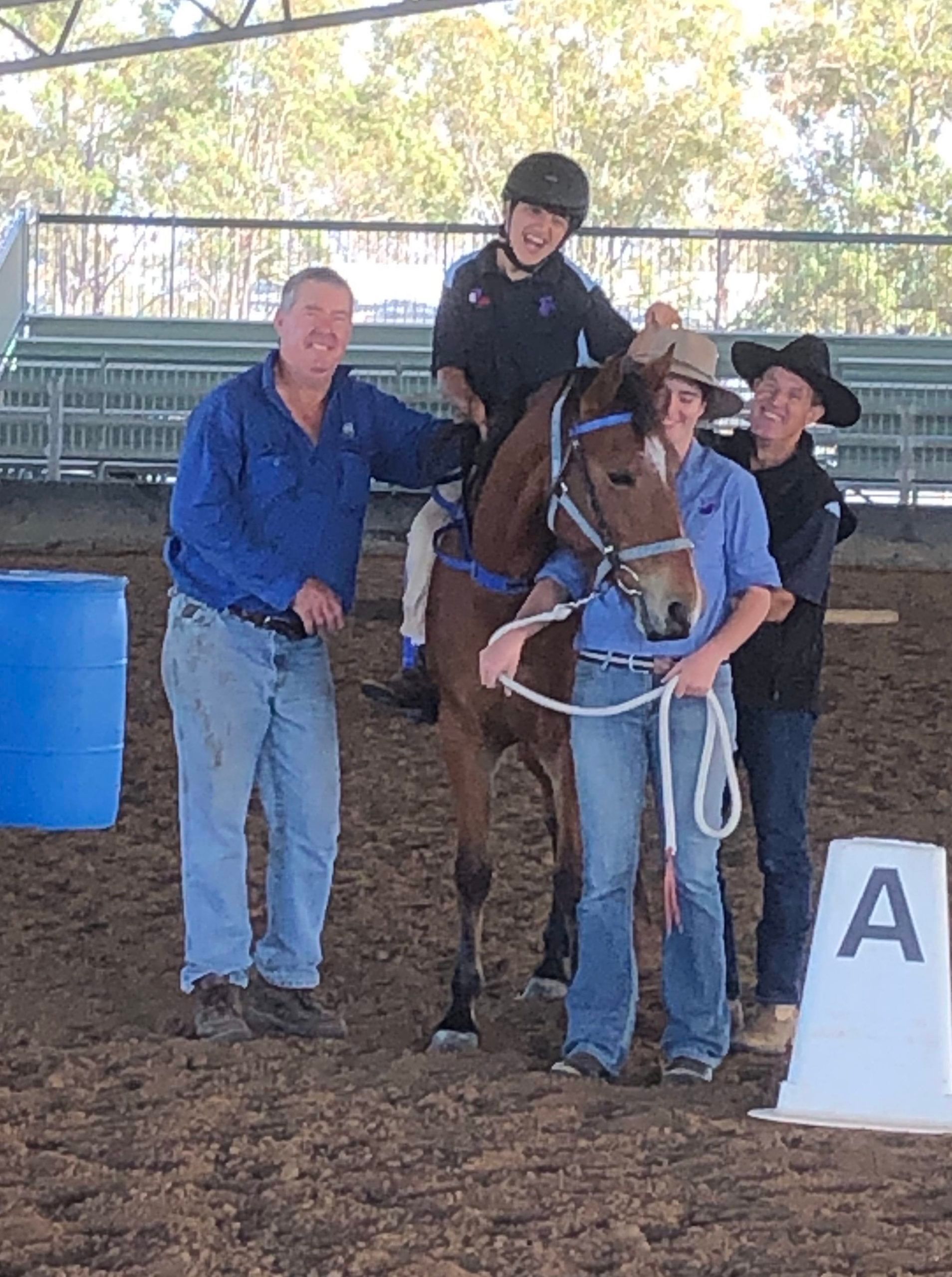 Four people with a horse in a riding arena. A person rides the horse. Three people smile, assisting.