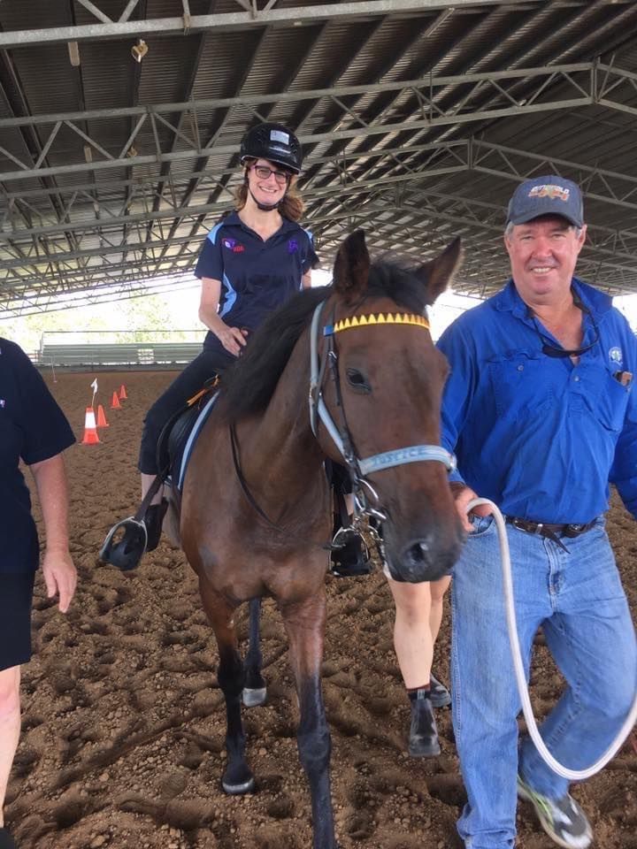 A woman riding a brown horse in an arena, led by a man. Another person is partially visible on the left.