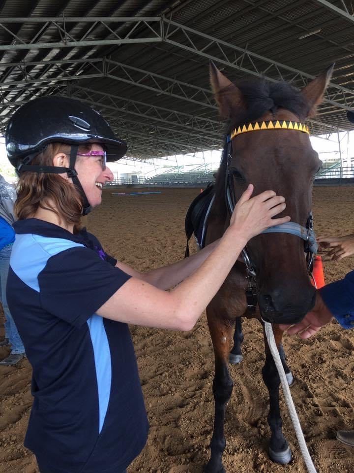 Woman in helmet and equestrian attire pets a brown horse in an arena.