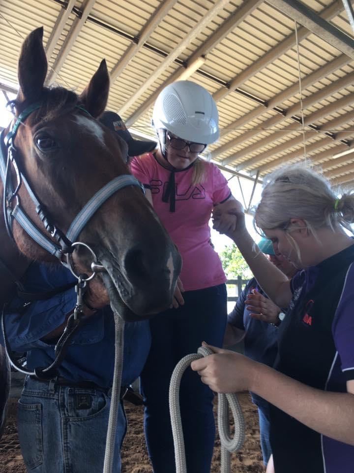 Woman in helmet adjusts horse's bridle as another holds a rope, all under a shelter.