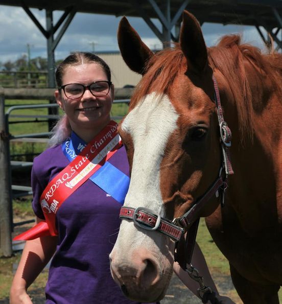 Person with glasses and a horse wearing a champion ribbon. Outdoor setting, horse is brown and white.
