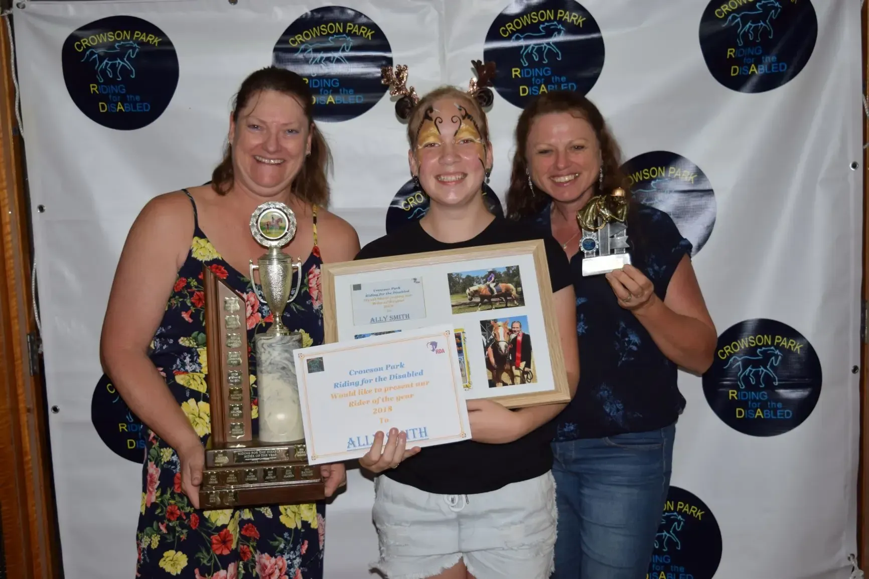 Three people smiling, holding trophies and certificates, against a banner with horse logos.