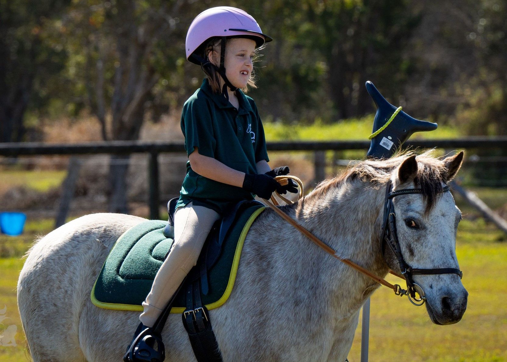 Girl in helmet riding a white pony outdoors, wearing green shirt and gloves.