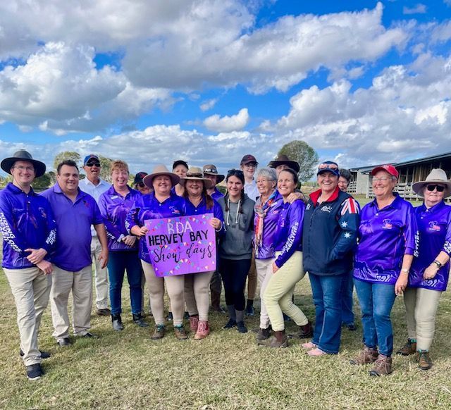 Group of people wearing purple shirts, holding a sign in an outdoor setting with a cloudy sky.