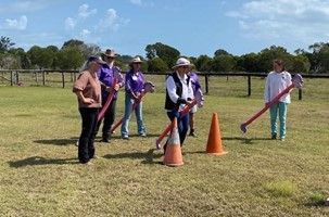 Group of people practicing with stick horses around orange cones on a grassy field.
