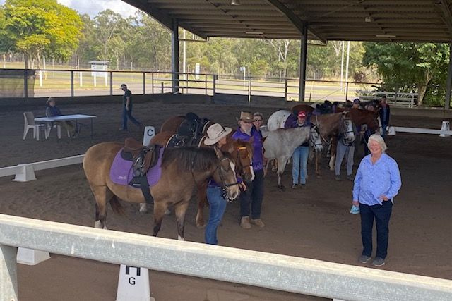 Horses with riders and handlers in an outdoor arena under a covered area; people watching in background.