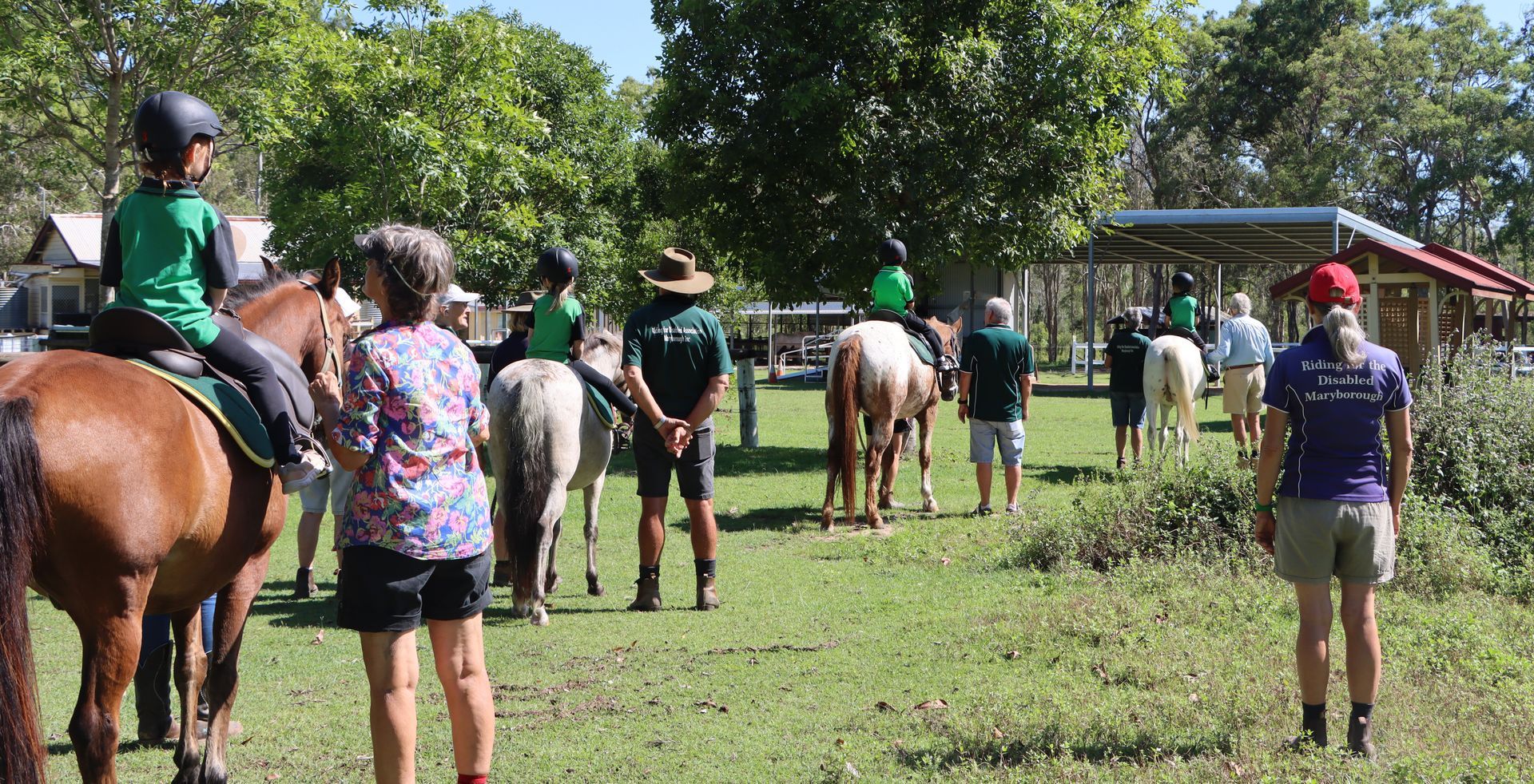 People on horseback and on foot in a grassy field. Riders wear helmets and trainers stand nearby.