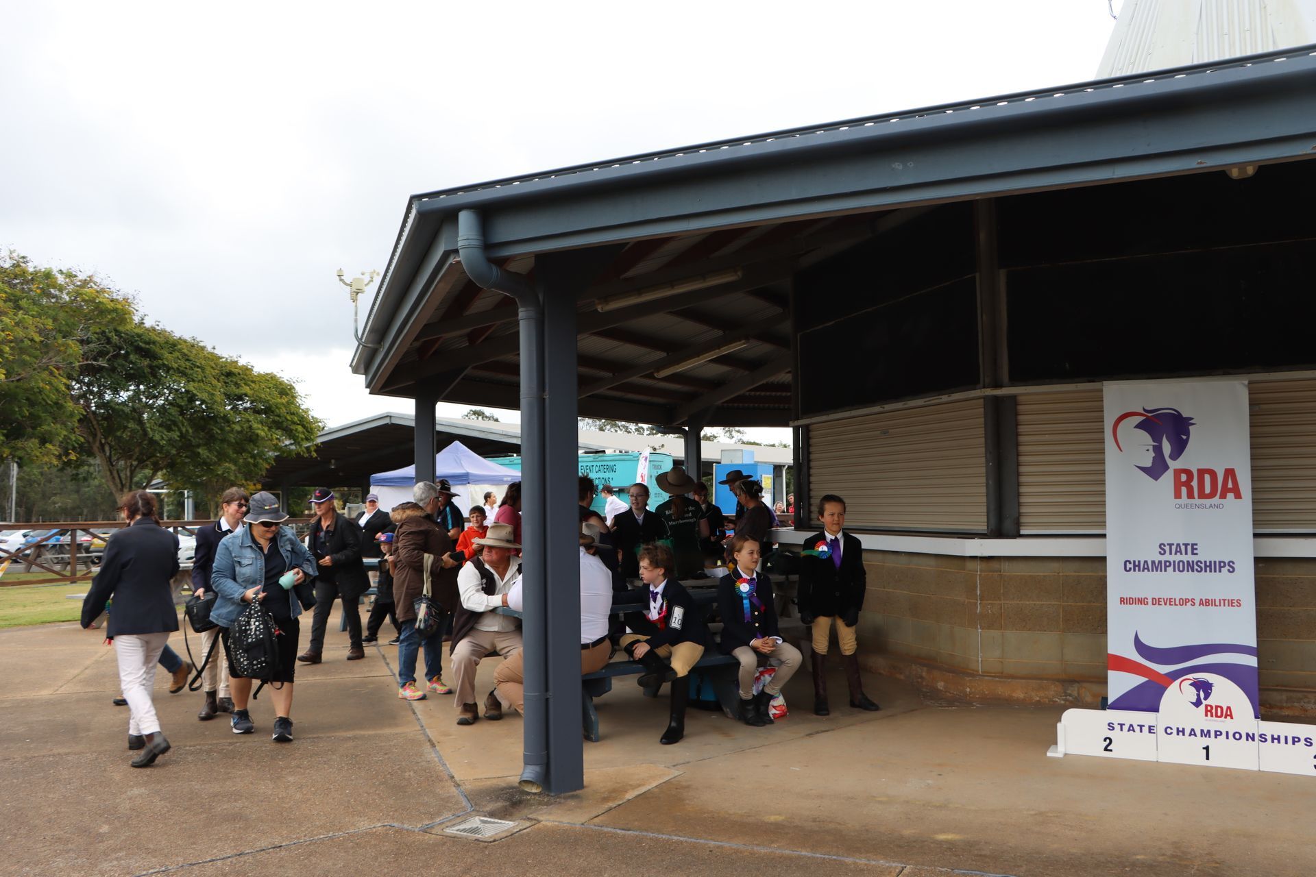People gathered under a covered area with an RDA banner at an outdoor event.