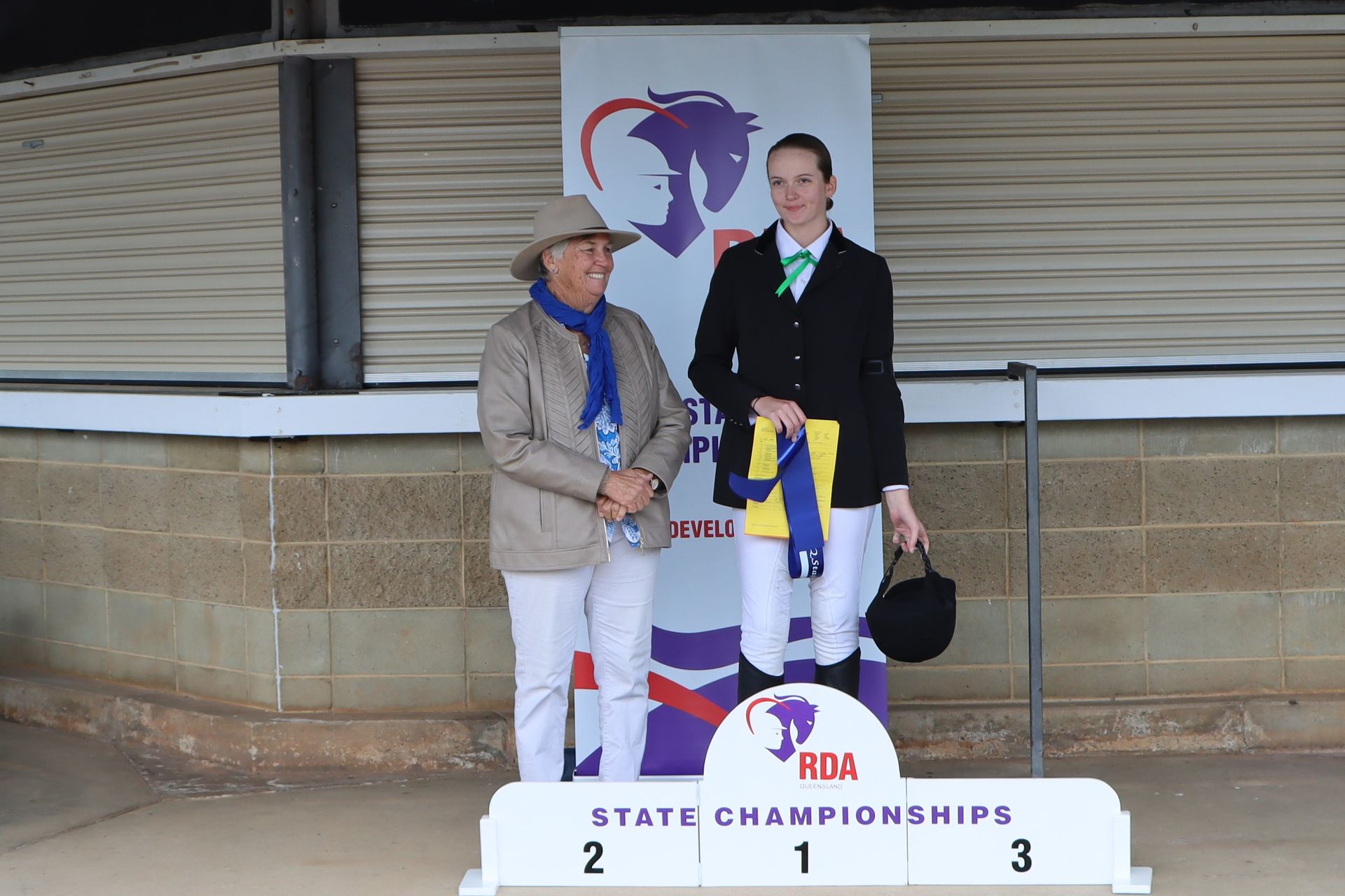 Two women on a podium at an equestrian event. The first place winner holds a riding helmet and a ribbon.
