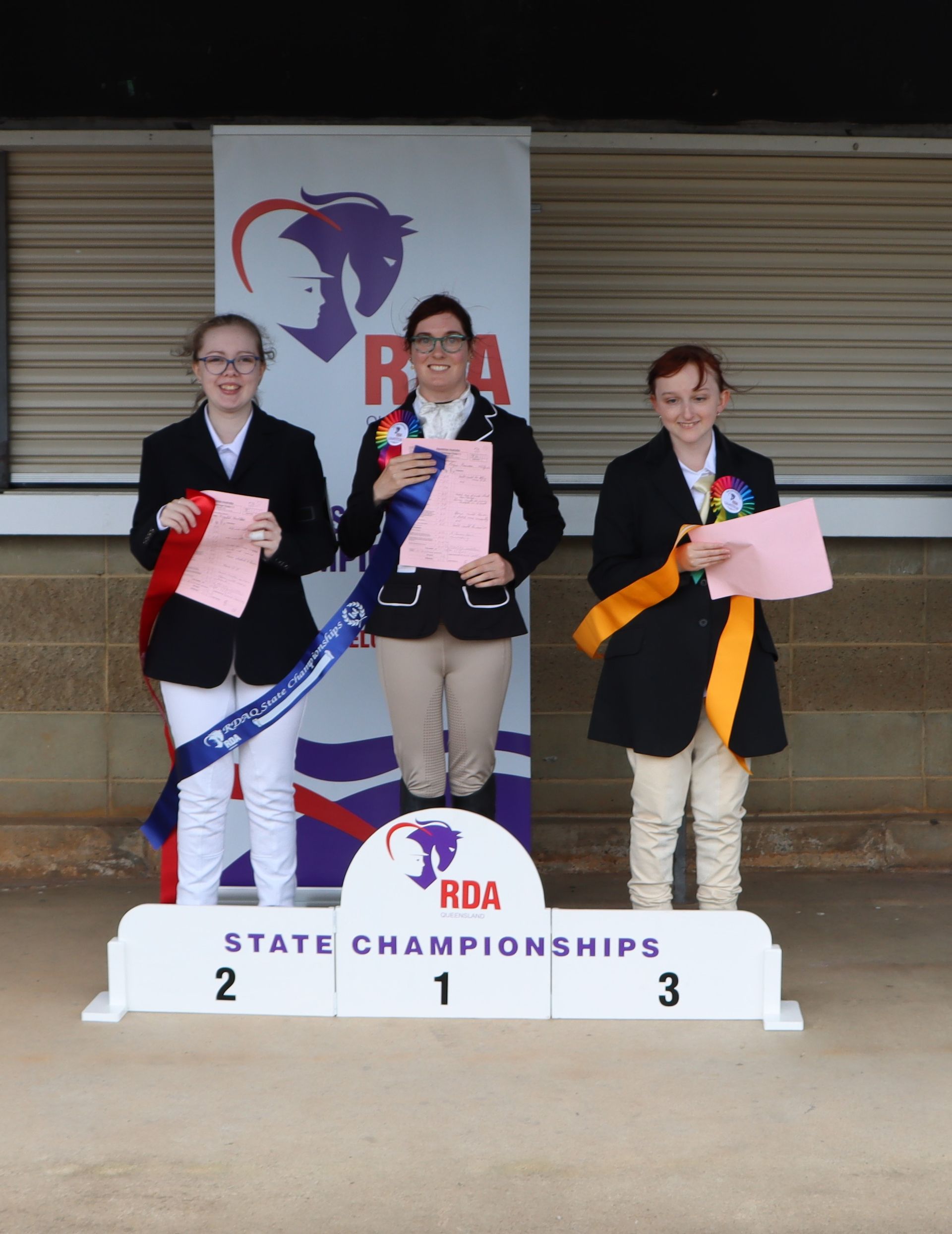Three riders stand on a podium with ribbons, celebrating at the RDA State Championships.