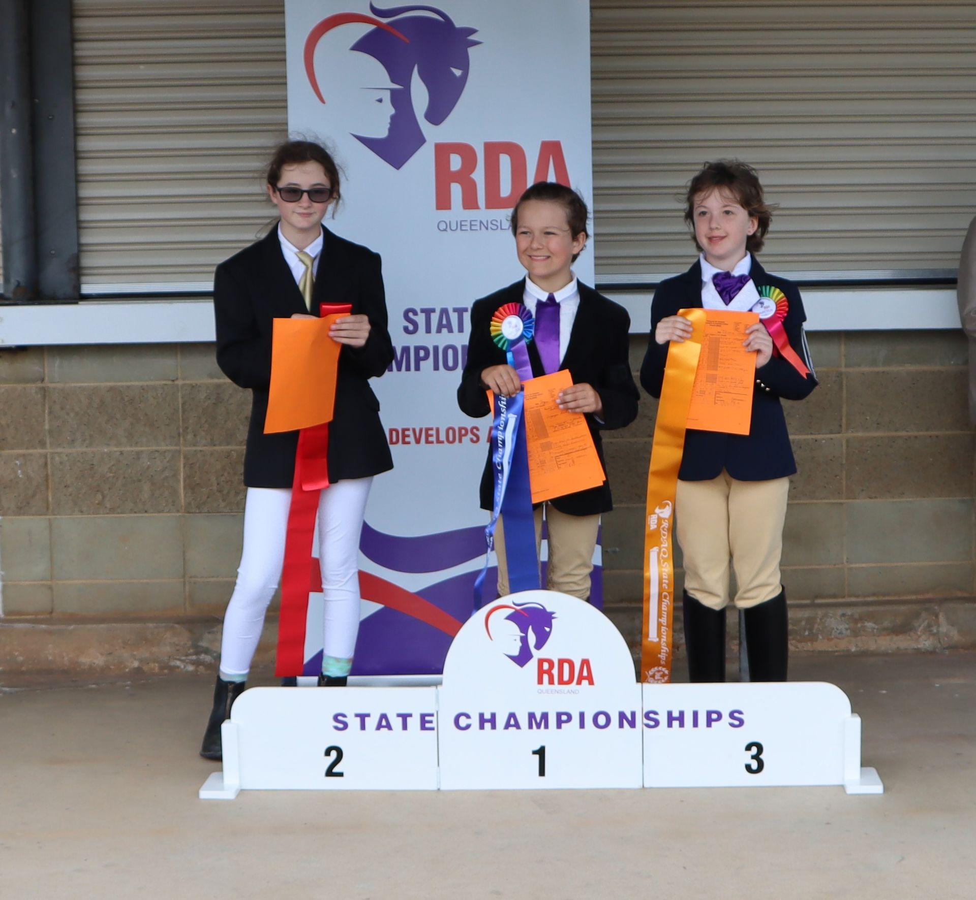 Three children on a podium holding ribbons. 