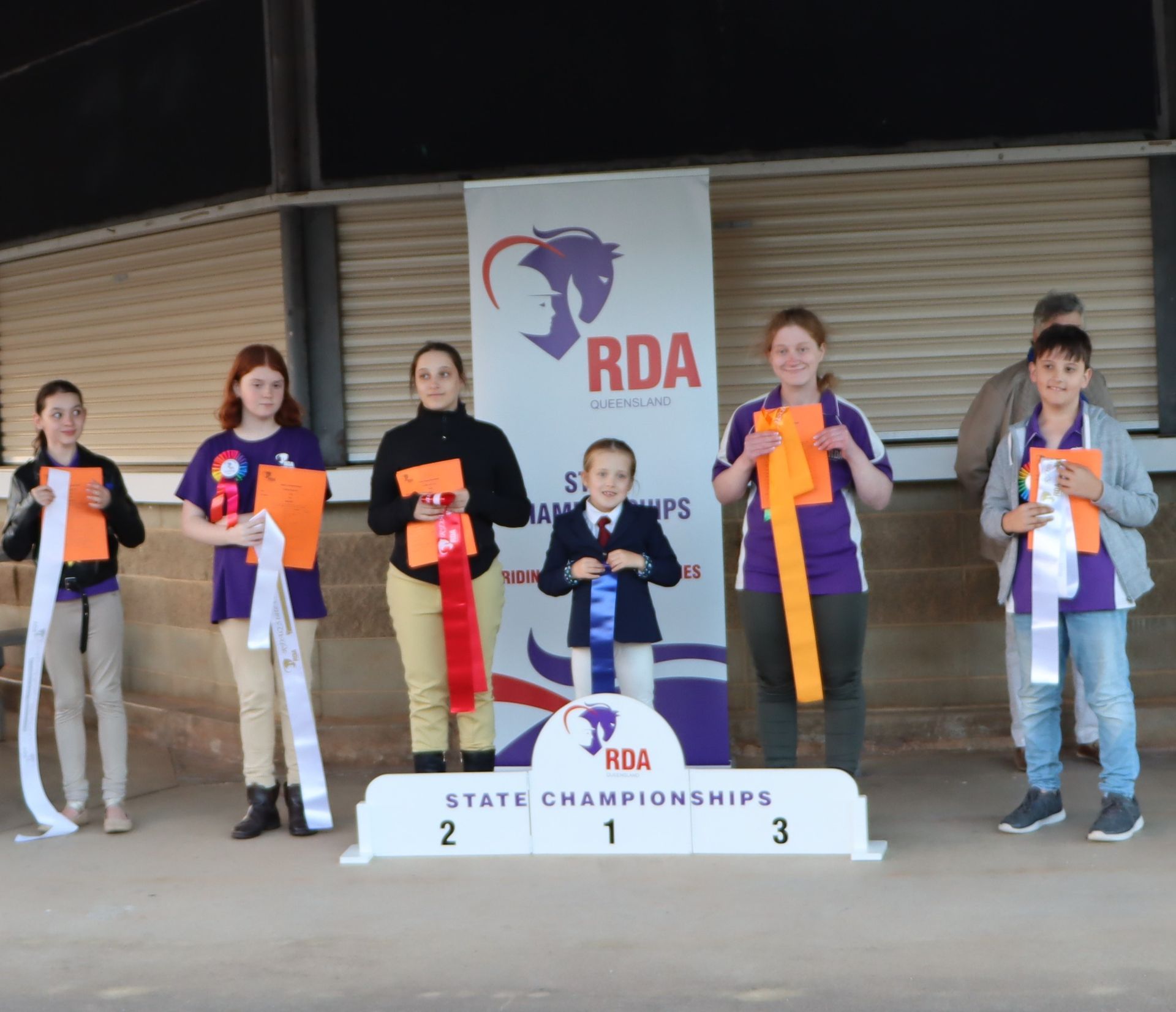 People on a podium holding ribbons. RDA State Championships banner in the background.