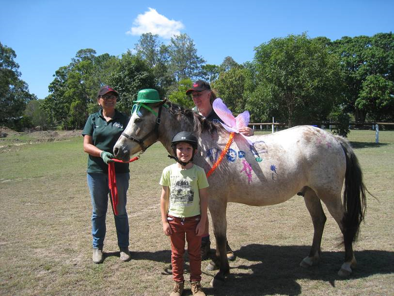 Woman, child, and horse outdoors; horse decorated, with hats, wings, and colorful paint.