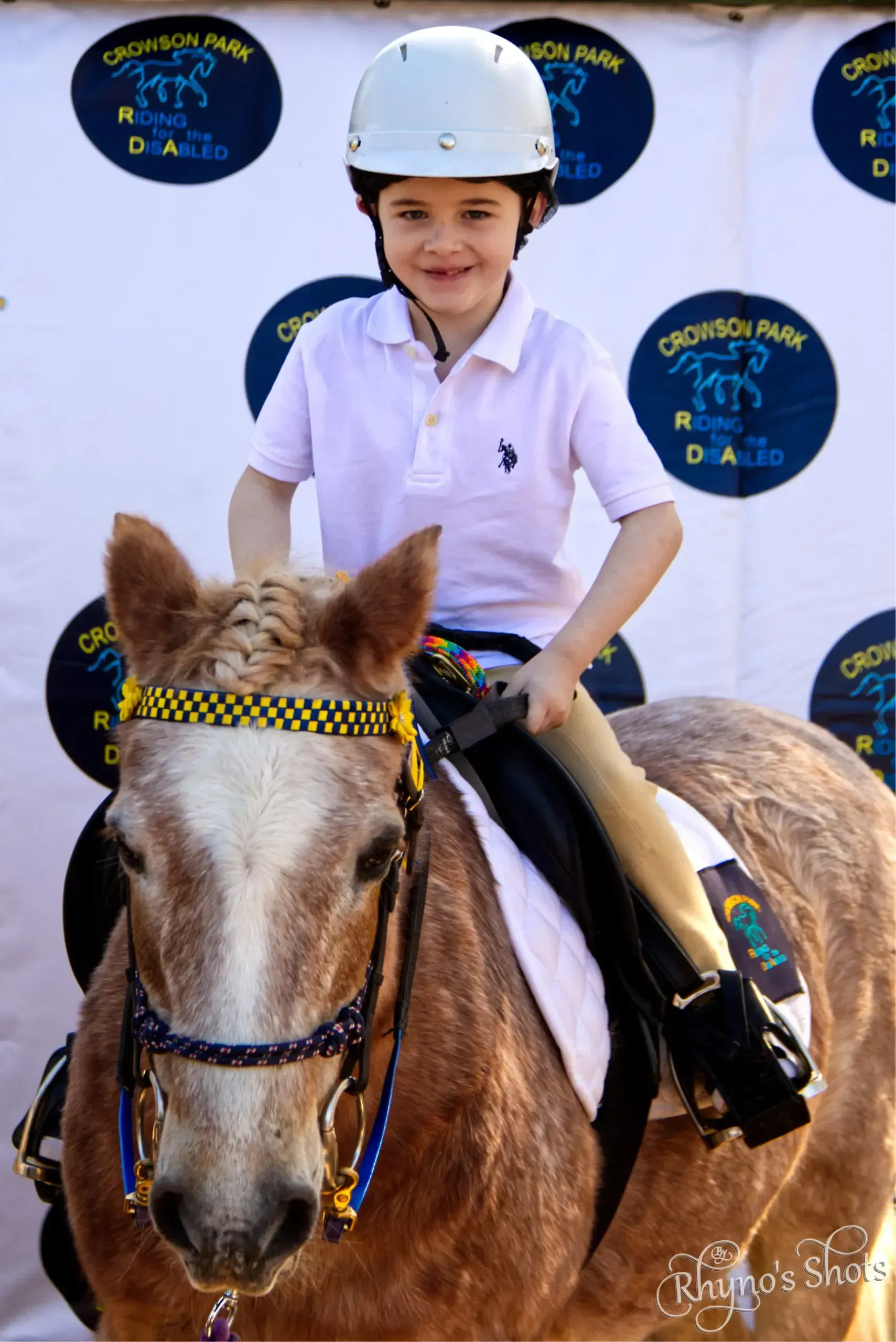 Young rider on a pony at a riding event, smiling, wearing a helmet and light pink shirt.