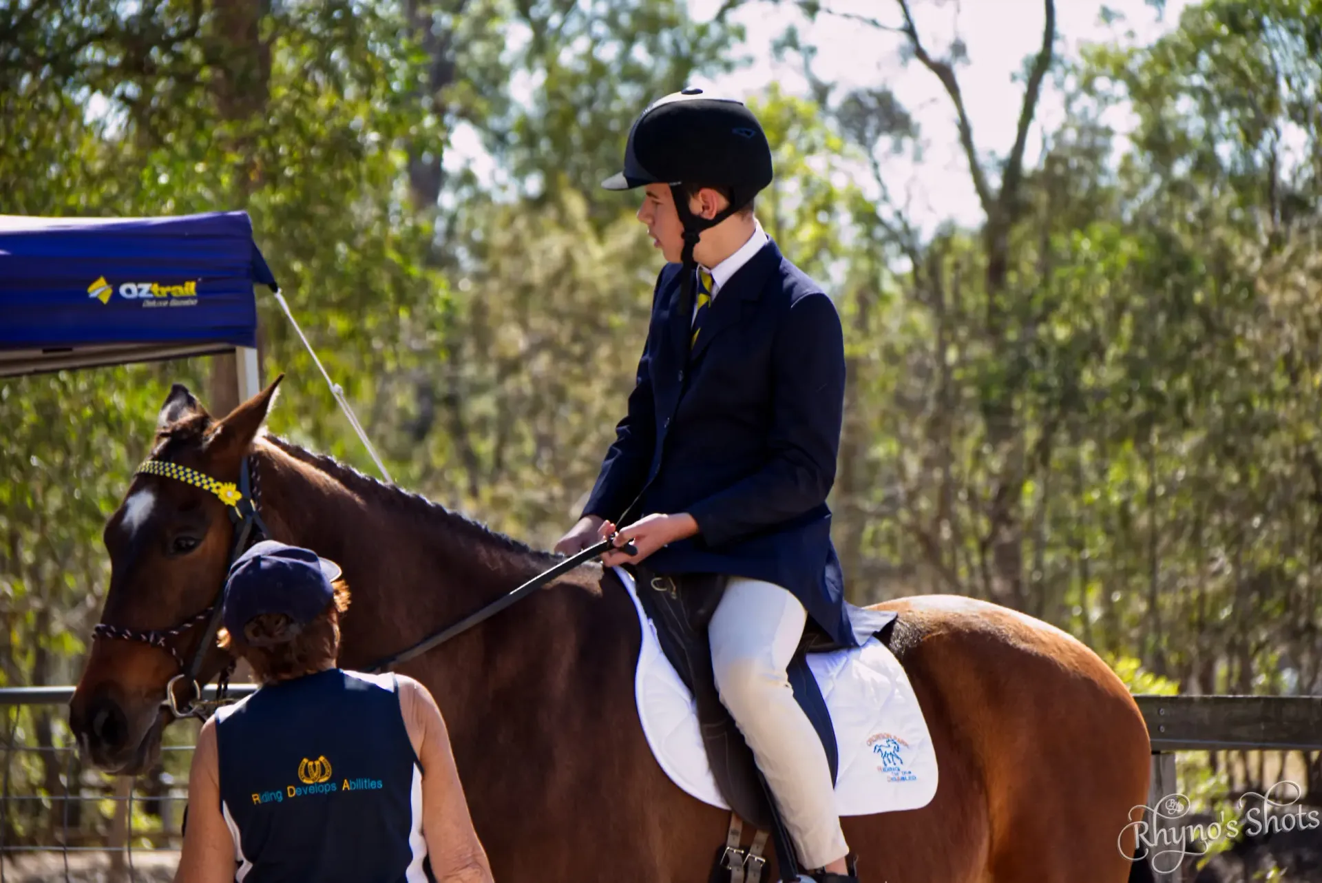 Person riding a brown horse in an outdoor arena, wearing formal riding attire. Another person stands nearby.