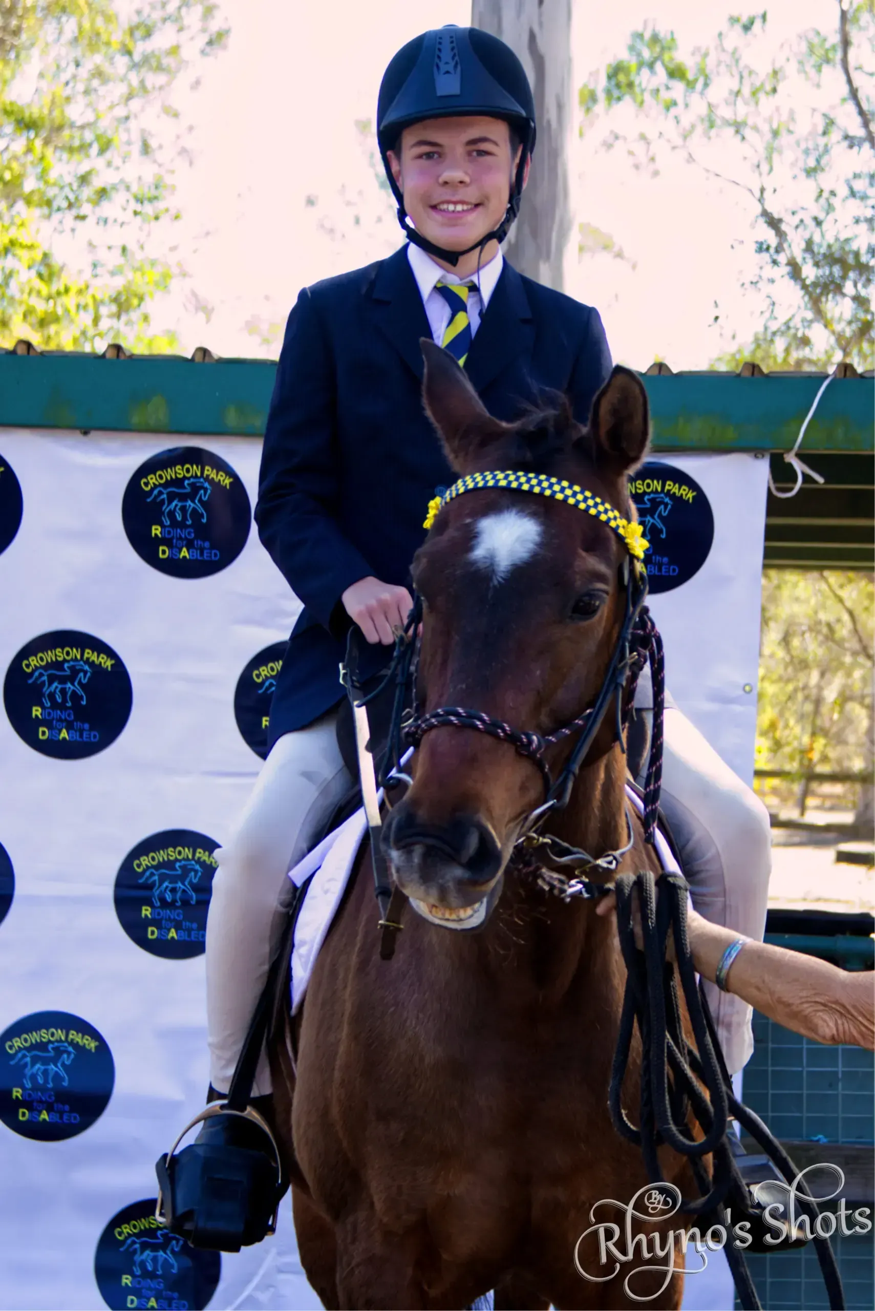 Young rider in blue blazer on a brown horse, with a ribbon, outdoors.