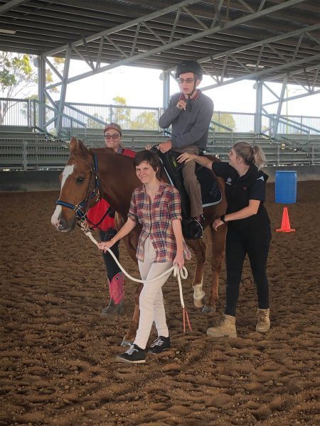 Person riding a horse with assistance from two adults inside a riding arena.
